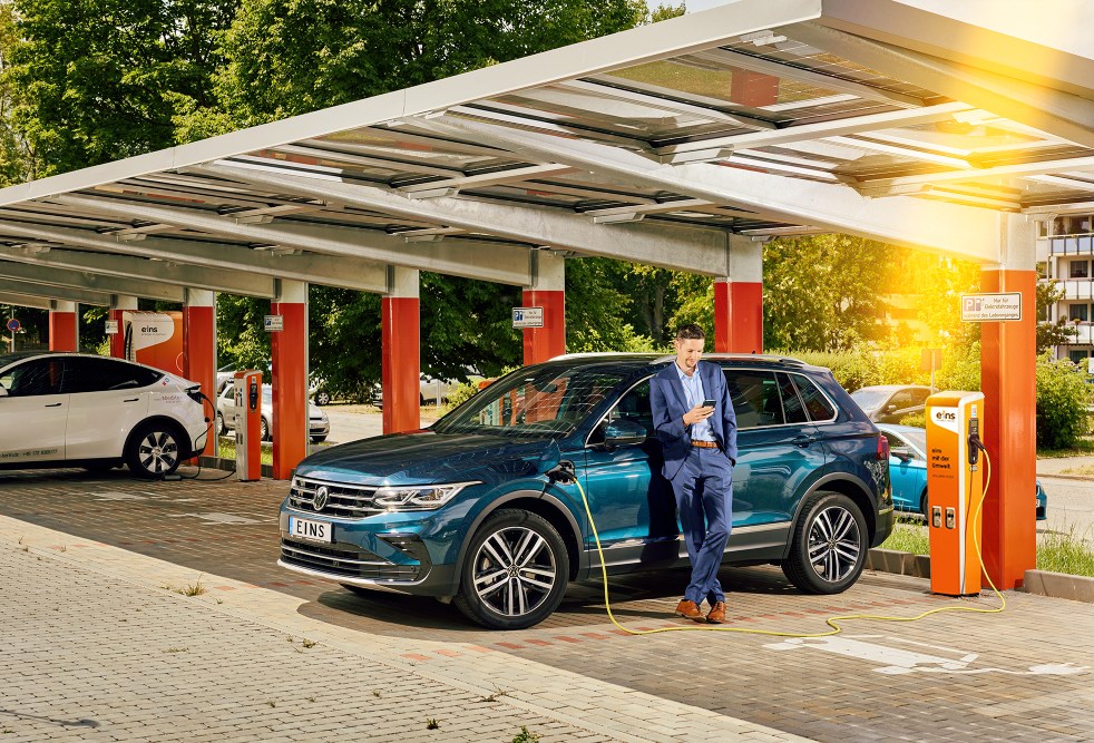 Geschäftsmann lehnt an E-Auto während dem Ladevorgang an einer Ladestation von eins