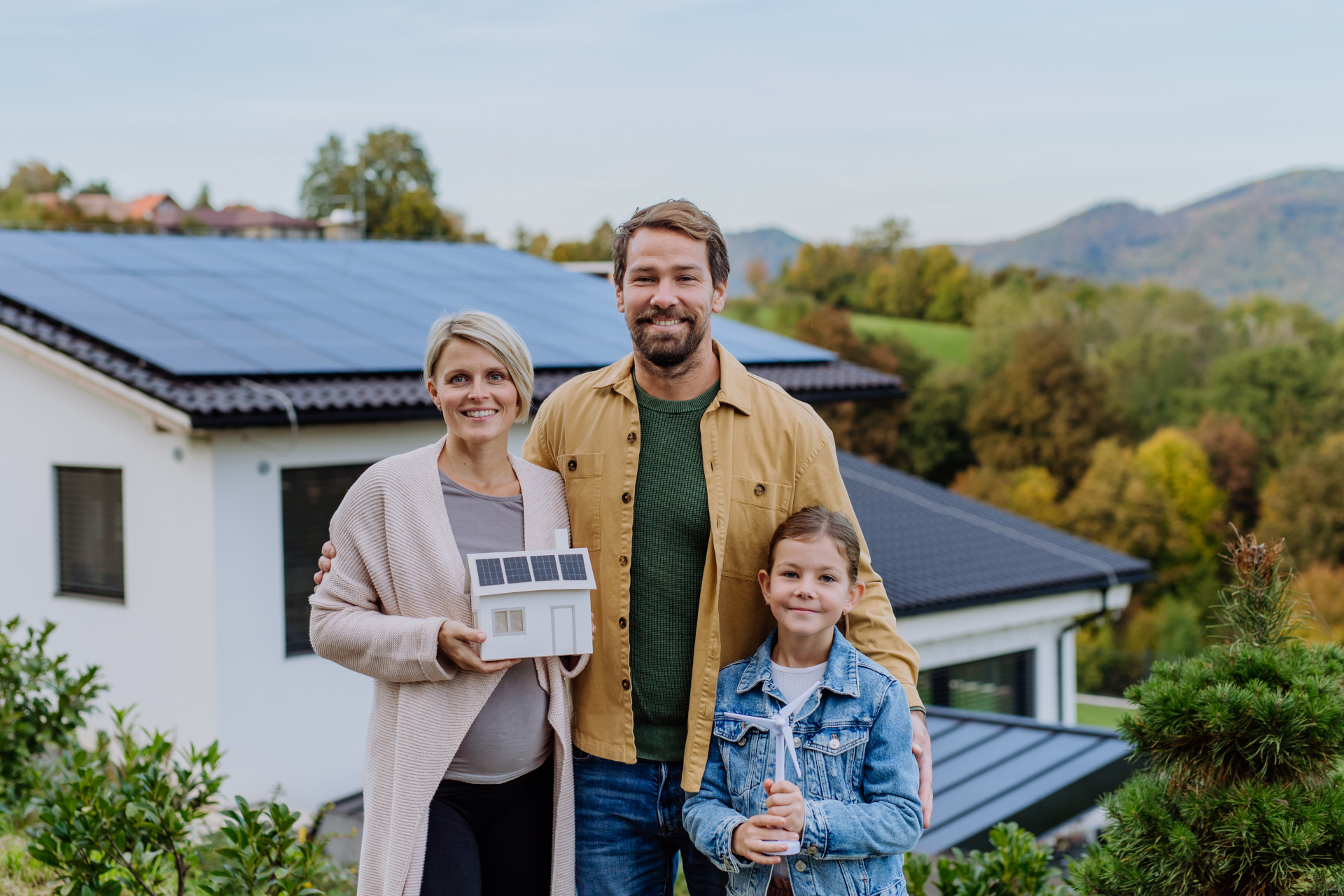 Vater, Mutter und Kind stehen vor ihrem Haus und schauen in die Kamera. Auf dem Dach des Hauses befindet sich eine Photovoltaikanlage.