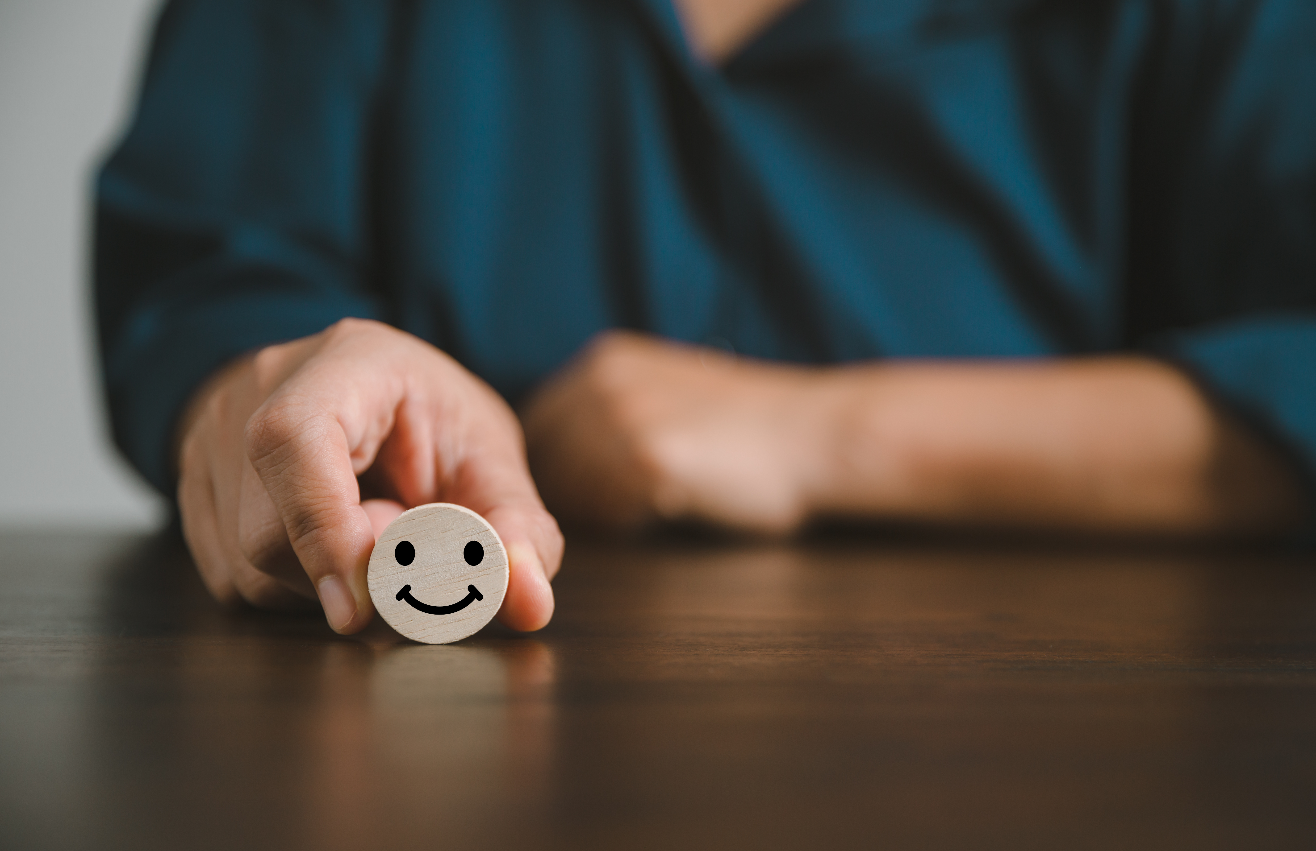 Mensch sitzt an einem Tisch und hält ein Smilie auf einem Holzstück in die Kamera