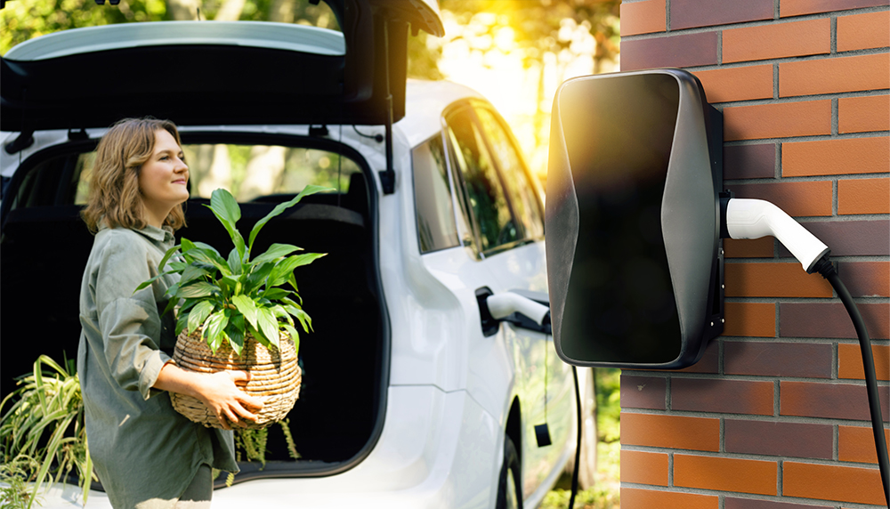 Frau räumte ihre Einkäufe aus dem Auto während man eine Wallbox im Vordergrund am Haus sieht