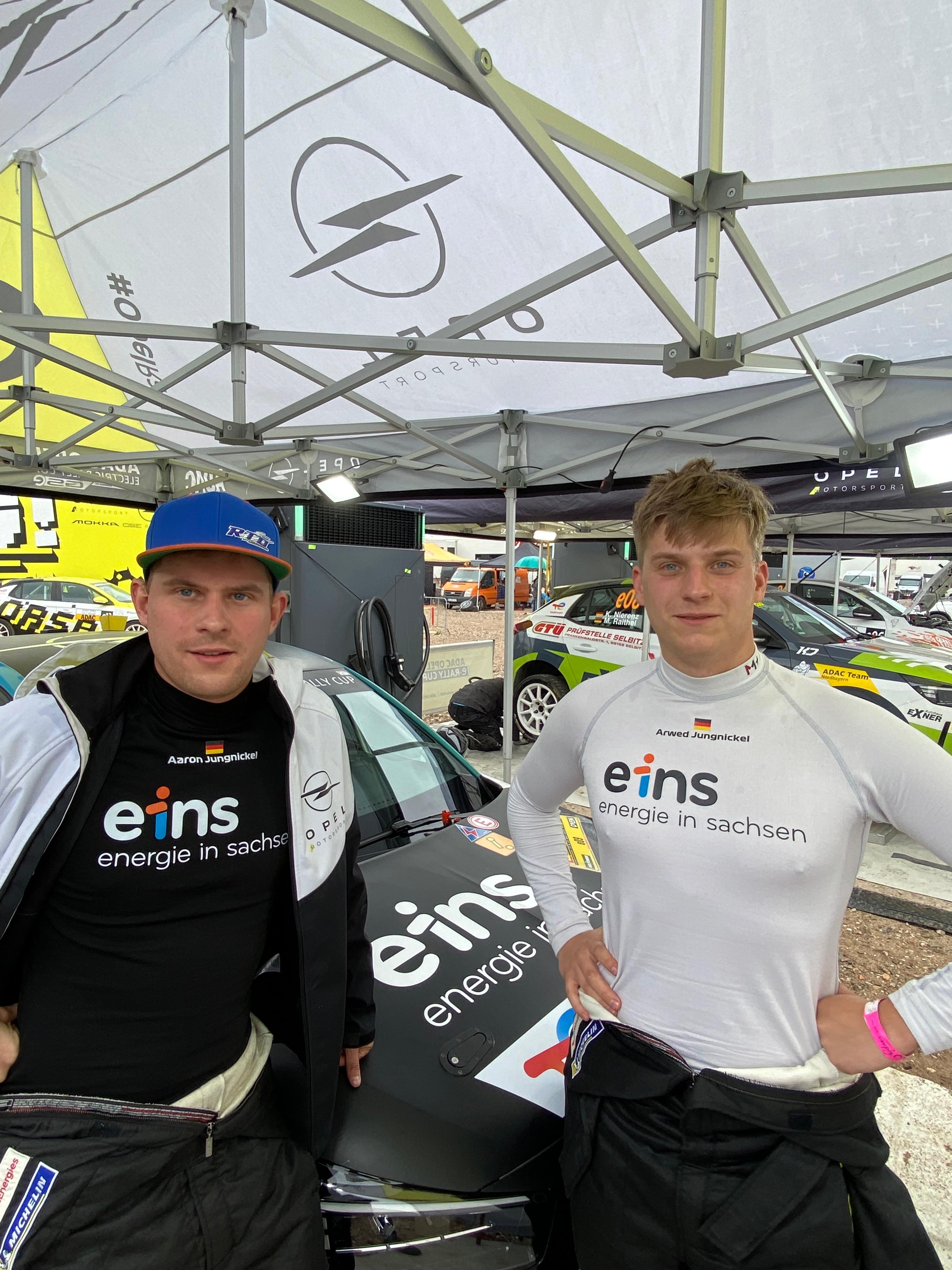 Aaron und Arwed stehen vor dem Rallye-Auto und tragen ihr eins-Trikot.