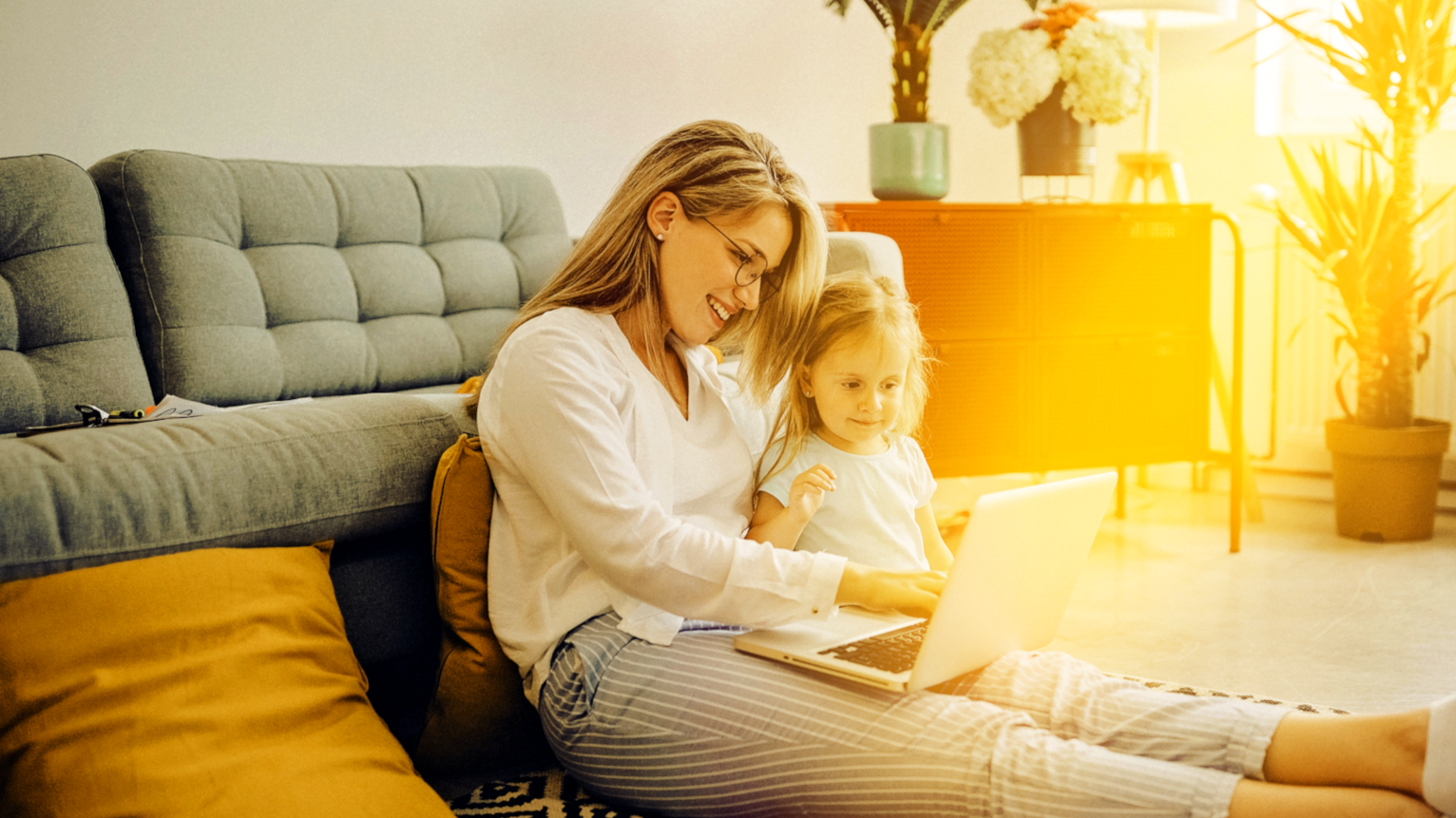 Mutter und kleine Tochter sitzen gemeinsam auf dem Teppich vor der Couch und schauen in den Laptop