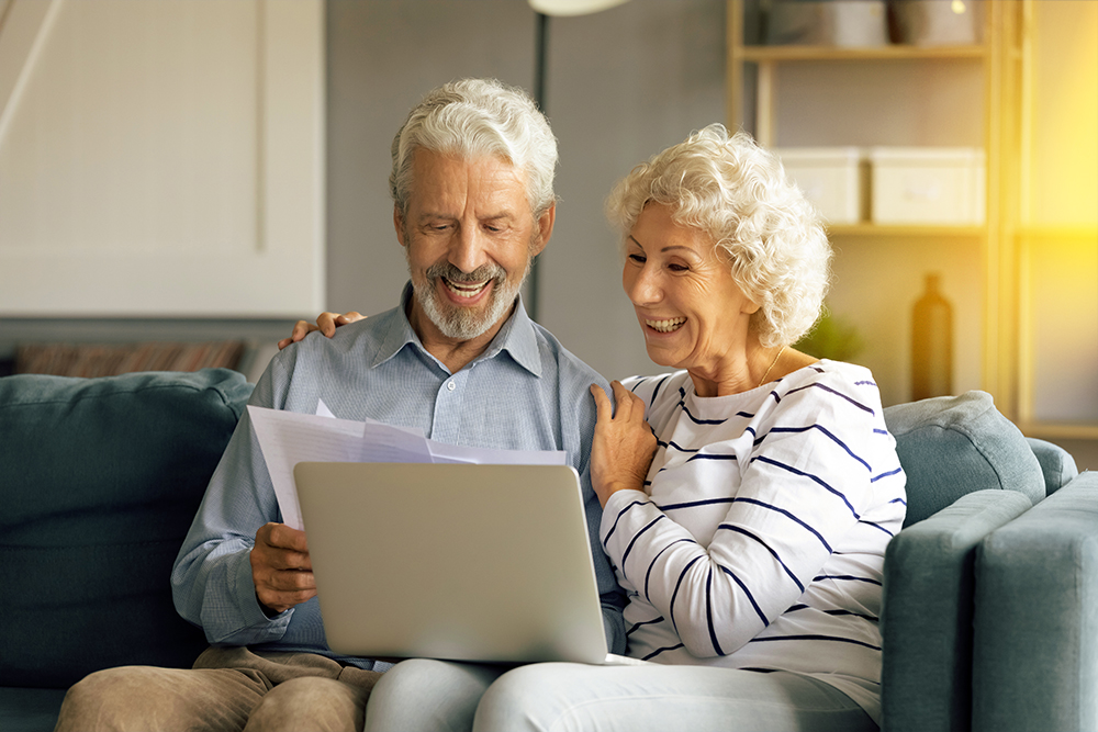 Rentnerpaar sitzt auf dem Sofa. Der Mann hält Dokument in seiner Hand, die Frau schaut auf den Laptop.
