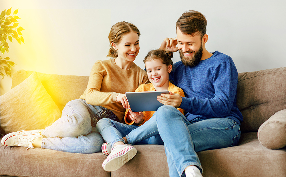 Eine Tochter sitzt mit ihren Eltern auf dem Sofa und sie schauen auf dem Tablet etwas an.