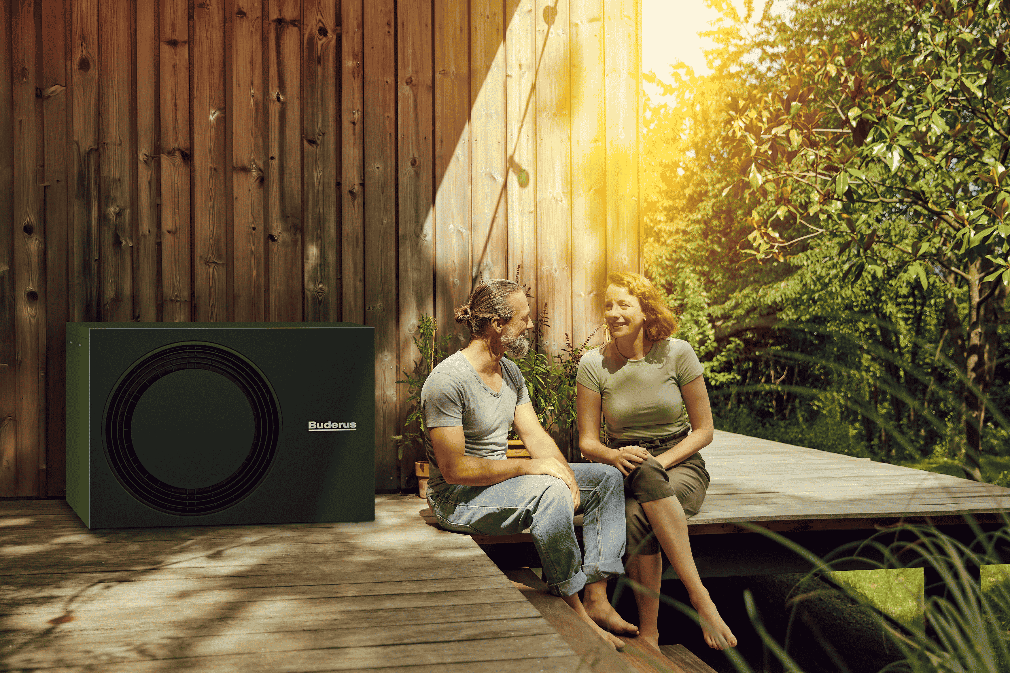 Ein Mann und eine Frau sitzen auf ihrer Holzterrasse vor ihrem Haus, neben ihnen steht eine Wärmepumpe von Bruderus