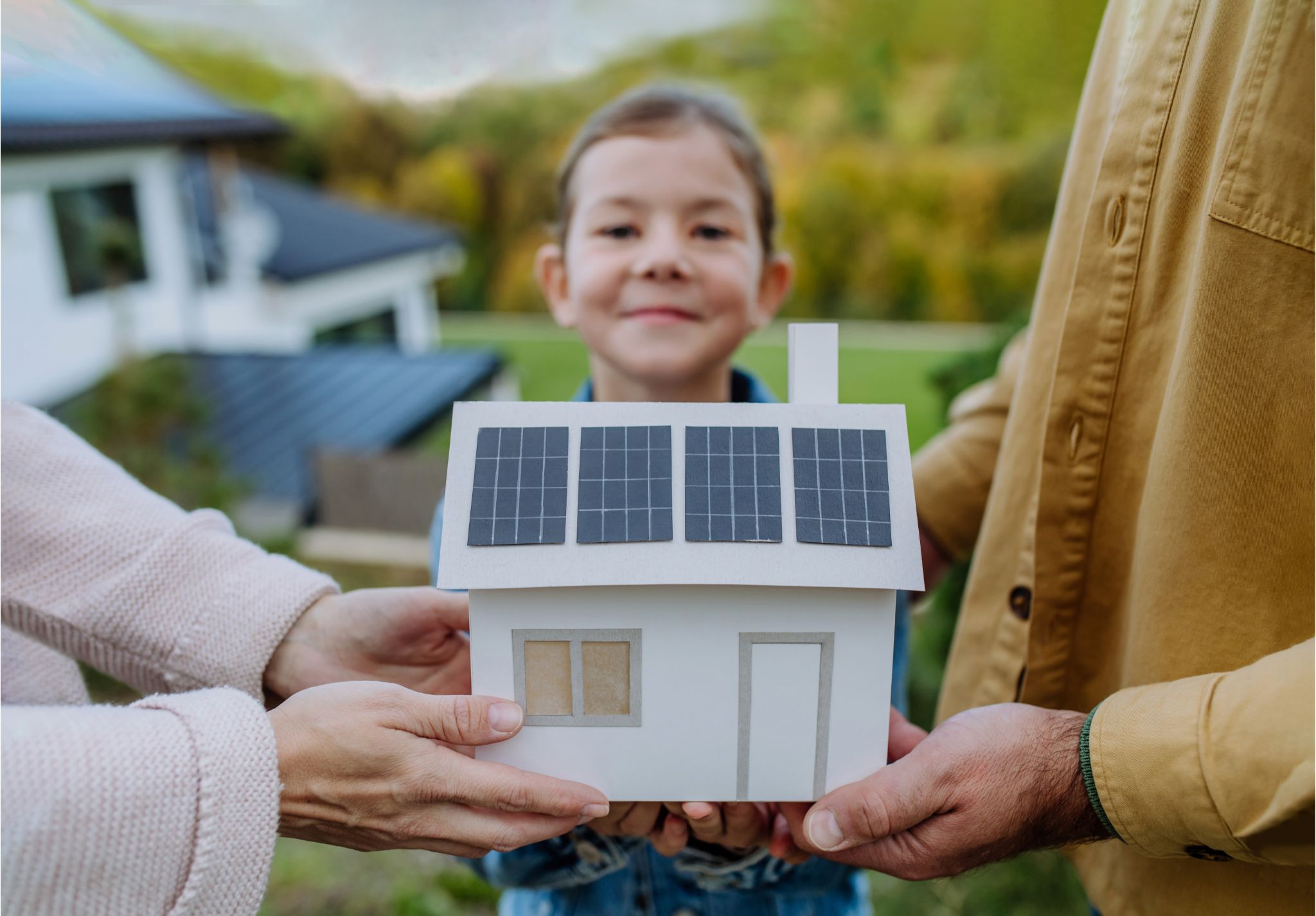 ein Mädchen mit stolzem Gesichtsausdruck hält gemeinsam mit ihren Eltern ein Papier-Hausmodell mit Solardach, welches symbolisch für ihr eigenes Einfamilienhaus steht 