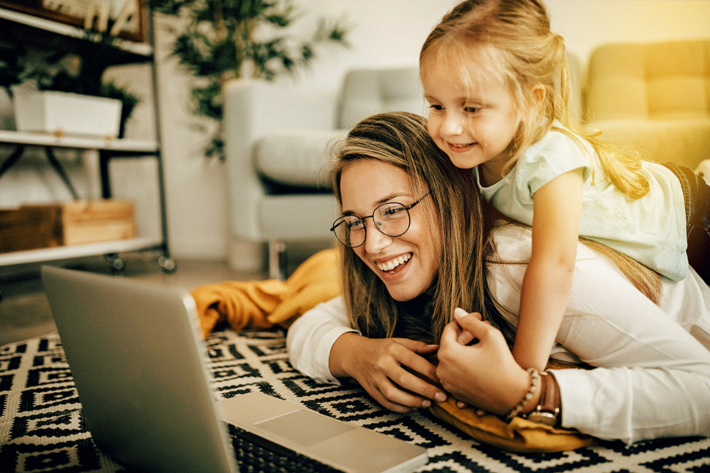 Mutter und kleine Tochter liegen auf dem Wohnzimmer-Teppich und schauen in den Laptop